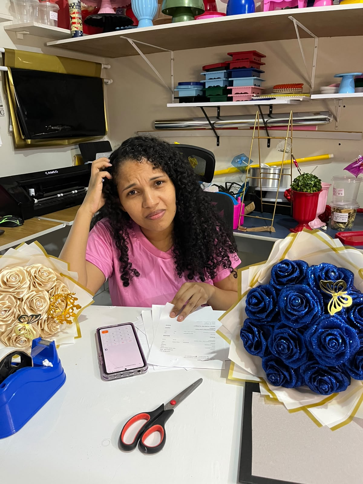 Hana conferindo as contas na bancada do ateliê, com celular e buquês artesanais